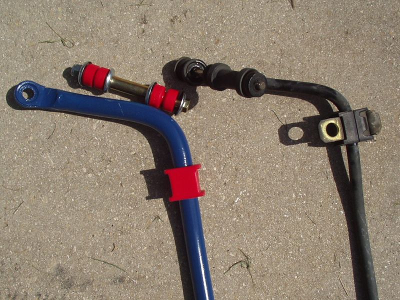 Blue 21 mm aftermarket sway bar with red polyurethane bushings and end links, laid next to the thinner black stock sway bar